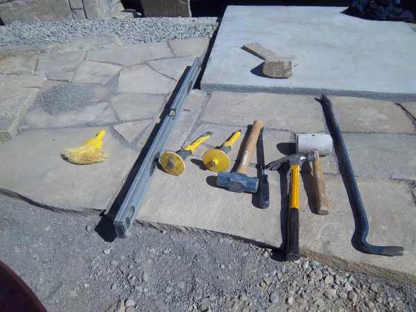 Tools for building a flagstone walkway. Whisk broom, 4 ft. level, 2 stone chisels, a 3 lb sledge hammer, a pry bar, a claw hammer, a rubber mallet (dead blow hammer) and a large pry bar.