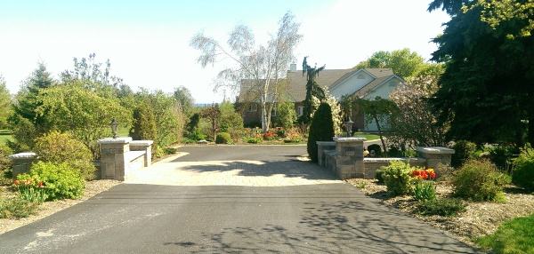 A Driveway entrance with block pillars connected with low block walls stands out with a band of brick pavers crossing through the asphalt.